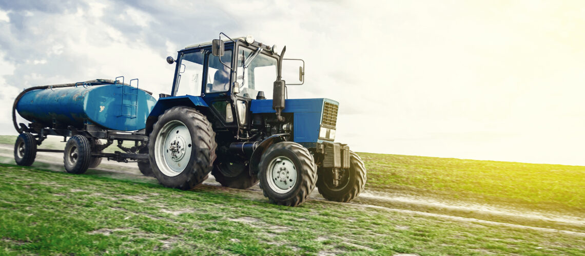A blue tractor with a barrel trailer rides along the spring field along the road.