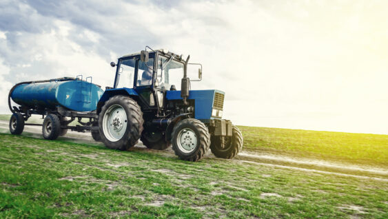 A blue tractor with a barrel trailer rides along the spring field along the road.