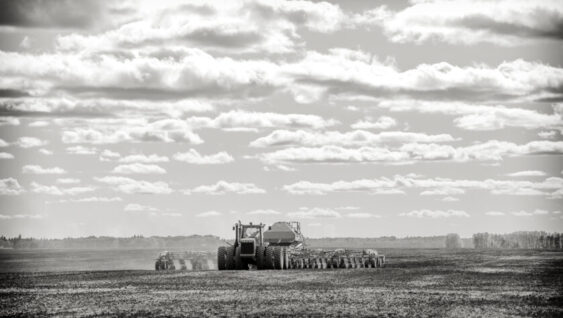 A tractor pulling a seeder and fertilizer tanks planting a crop in a black and white countryside summer landscape.