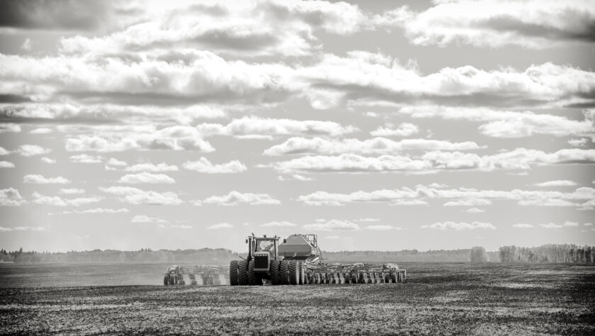 A tractor pulling a seeder and fertilizer tanks planting a crop in a black and white countryside summer landscape.