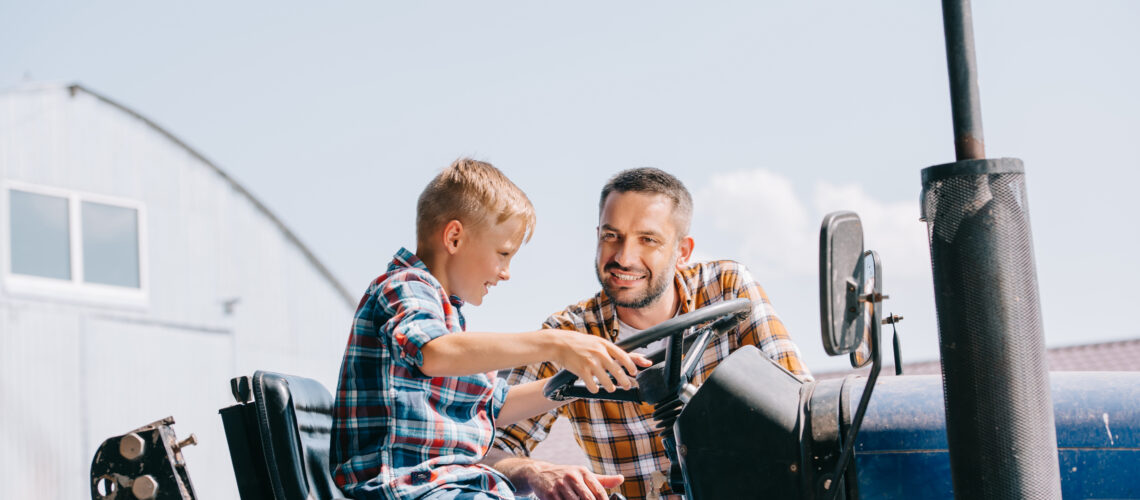 A happy father looking at his smiling son sitting in a tractor on farm.